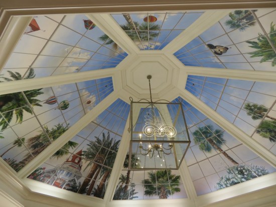 Circular ceiling fresco divided into eight wedges with painted palm trees, blue skies, clouds, and distant riding balloons, one of which is shaped like Mickey Mouse's head.