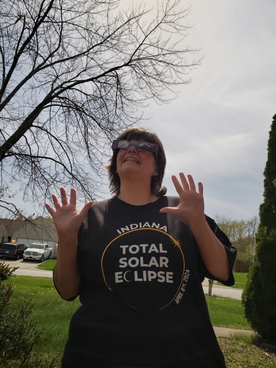 Anne outside wearing eclipse glasses that say "North American Eclipse" on them and a black T-shirt reading "Indiana Total Solar Eclipse, April 8, 2024".