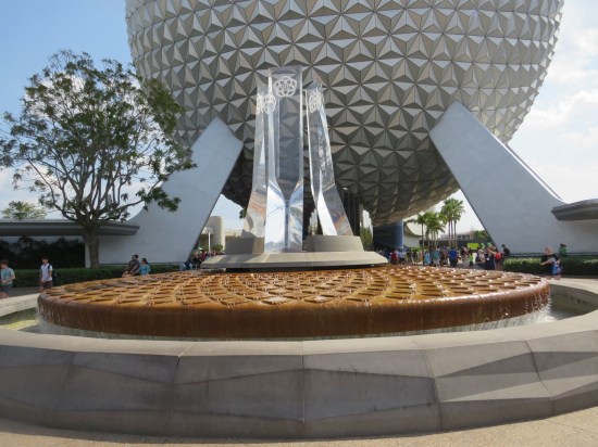 Four glass pylons with a Disney World symbol atop each. They stand atop a fountain that's a brown platform with crosscuts in it. Concrete seating surrounds the fountain. Spaceship Earth looms large in the background.