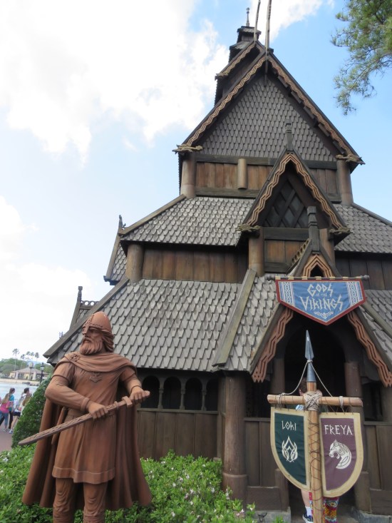 Tall Scandinavian building with one sign proclaiming "Gods of the Vikings" and a post with dual signs for Loki and Freya. Out front is a brown swordsman statue.