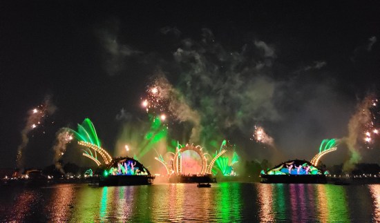 wide shot, green fountains, gateway screens now show inscrutable dancing figures. Lights reflect as stripes across the water.