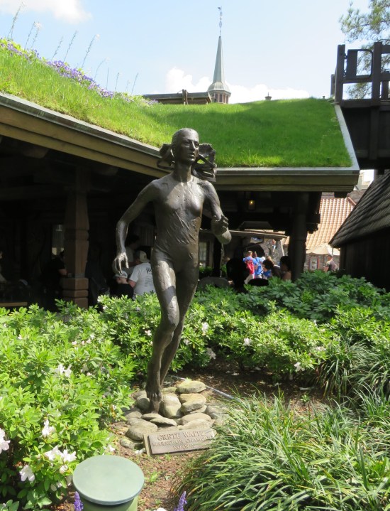 Statue of a female runner standing amid a bunch of flowers and greenery behind a building whose roof is also covered in greenery.
