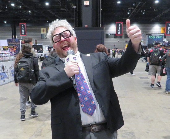 Cosplay: old loudmouth with white hair, cheap suit and purple tie, pretending to bellow into microphone.