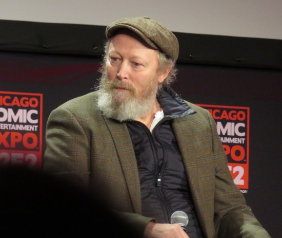 Lars Mikkelsen sitting thoughtfully with a mic, wearing a nice hat and jacket.