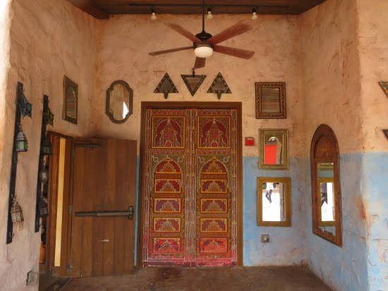 A locked wooden door with faintly Islamic trimmings, next to an open, unadorned utility closet door. Mirrors, triangular hangings, and a ceiling fan adorn this stone alcove.