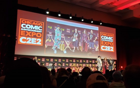 Three people and an emcee onstage, as viewed from several rows back in darkness. Large projection screen shows them more closely but at an awkward because we're sitting over at stage right.