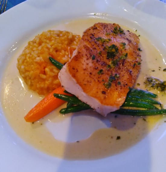 Fish, risotto, green beans and one carrot slice on a plate ringed with butter.