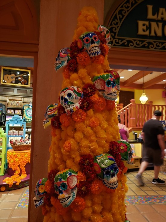 Orange leis arranged in a Christmas tree shape with painted skulls hanging on it. It sits in an indoor gift shop.