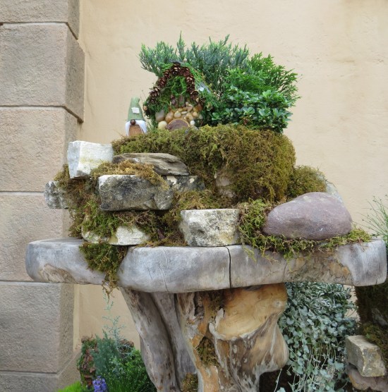 Tiny house with roof made of evergreen branches, sitting on a moss bedding atop a stone pedestal on the side of a full-size beige building.