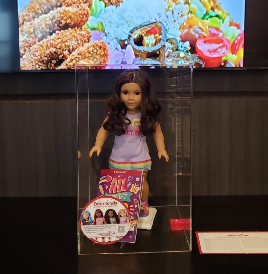 A single American Girl doll in a glass case atop a hotel lobby end table. Above it, a wall-mounted TV shows pics of the hotel restaurant's fare.