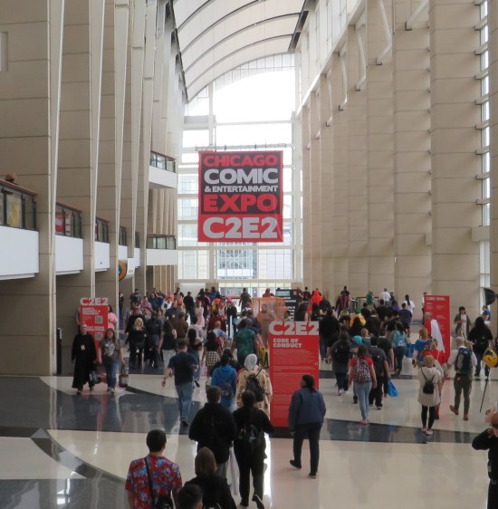 Giant C2E2 banner hanging from vaulted white convention center ceiling. Fans come and go around us.