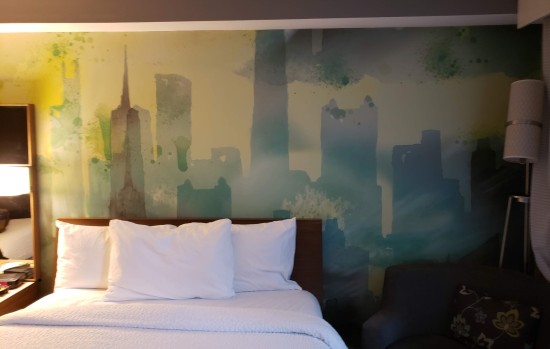 Shaky silhouette mural of Chicago skyline in green under a yellow sky with one looming, dark green cloud. Hotel bed, chair and lamp obscure the bottom.