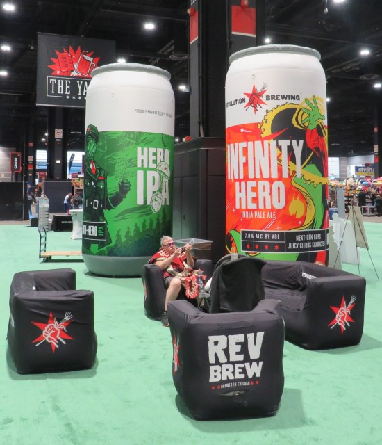 Two giant inflatable beer cans on green carpet next to some black puffy furniture.