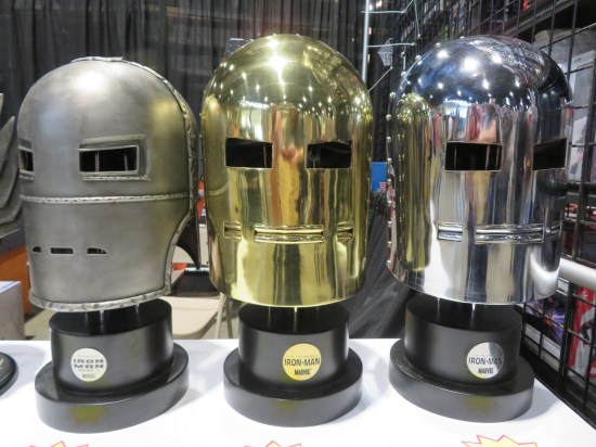 Table holding one dull metal Iron Man helmet, a shiny metallic one, and a shiny gold one.