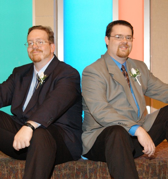 Two guys in suit jackets and ties sitting on a carpeted stage. The back wall has thin beige and blue glass panels alternating within white borders.