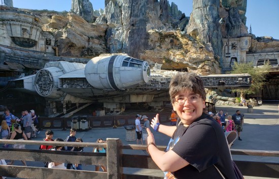 My wife gesturing wildly at the life-size Millennium Falcon parked far behind her. In the background are massive canyon walls. A few other tourists mill about.