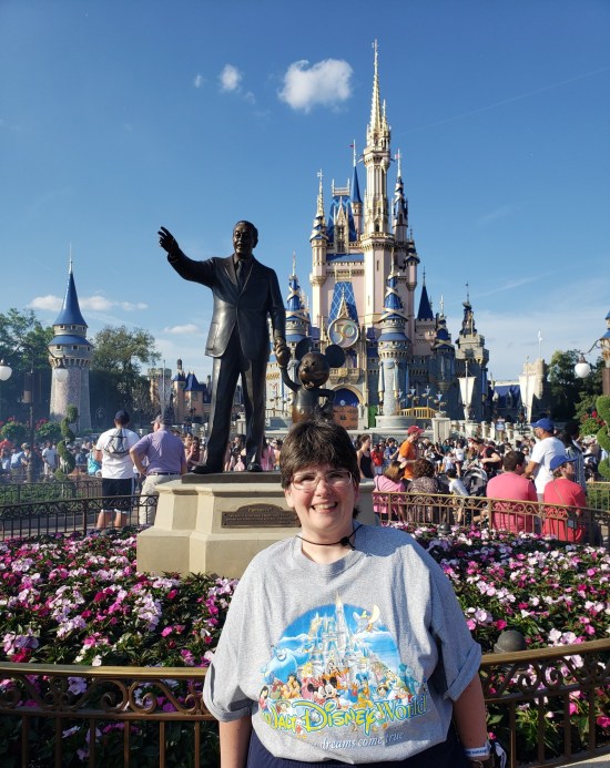 Anne in mouse-ears hat and Disney World T-shirt standing in front of Walt Disney statue, in turn standing in front of distant castle. A flower garden surrounds the statue; hundreds of guests surround the castle.