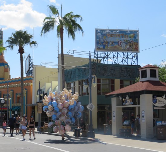 Streetside vendor selling blue, silver, and peach Disney balloons. On a roof is a fake billboard for an old-timey beach called Paradise Pier.