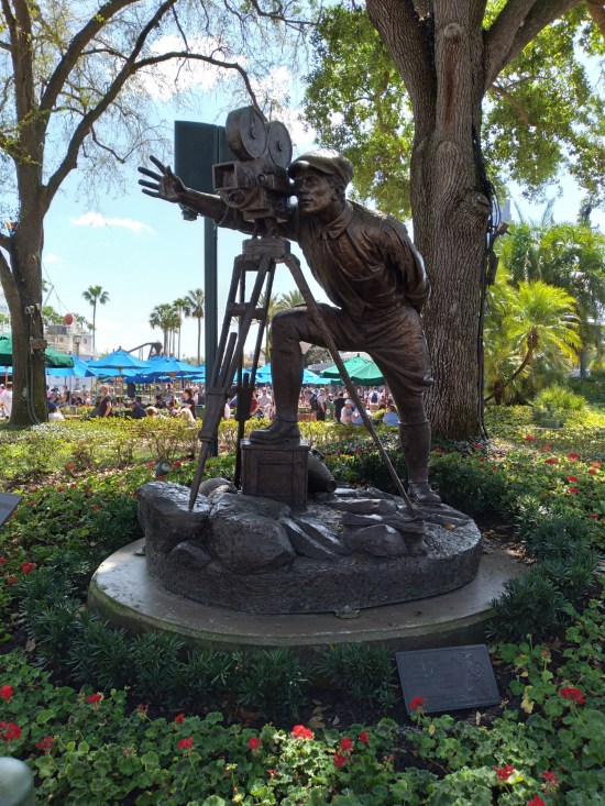 Bronze statue of a director resembling Billy Wilder, looking through a camera lens and waving with his free hand.