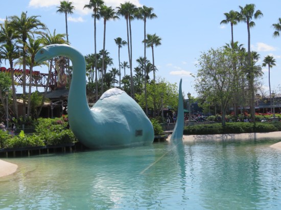 Big apatosaurus statue in a pond ringed by bushes and palms.