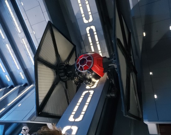 Half-size red-and-black TIE Fighter hanging on a blue wall, which angles at 45 degrees before the top half doubles back toward the ceiling.