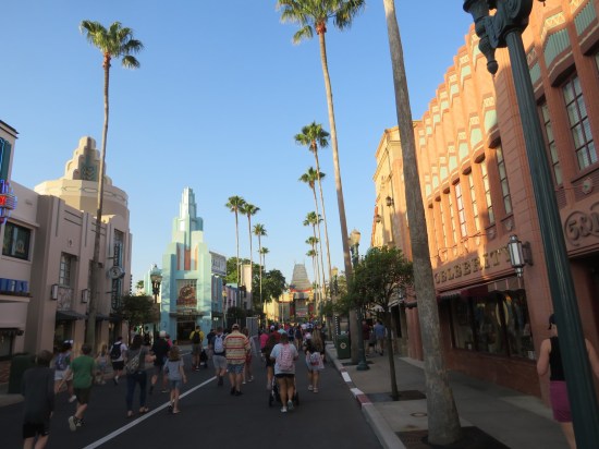 Tons of guests walking quickly down the theme park streets. Palm trees and gift shops line both sides.