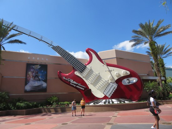 Red and white giant guitar partly buried on one side like a shovel jammed into the ground. Square building behind it has an Aerosmith poster, but almost no people around.