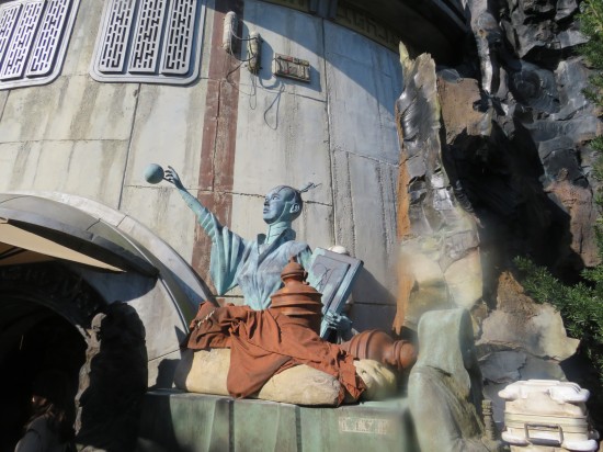 Blue statue of a bald woman with no pupils, holding a big tome in one arm and pointing at a floating ball with her free hand. Tan urns sit or lie around her on her concrete pedestal.