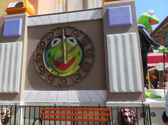 On the side of a colorful exterior hodgepodge wall is a large clock with Kermit's face in the middle and twelve circled stars instead of numbers. The hands point to 2:00.