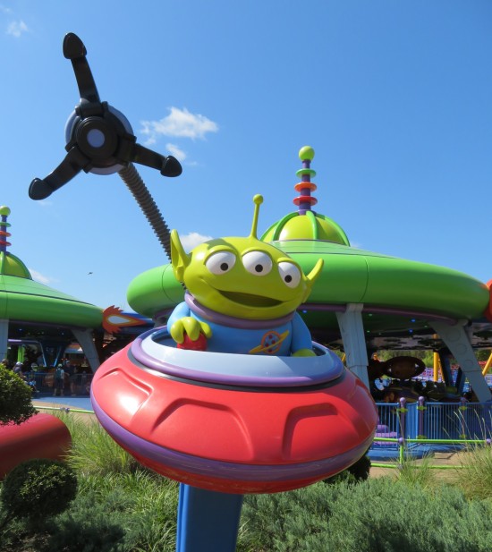 Little Green Man statue in a flying saucer. The Claw extends over one shoulder toward the camera. In the distance are kiddie rides.