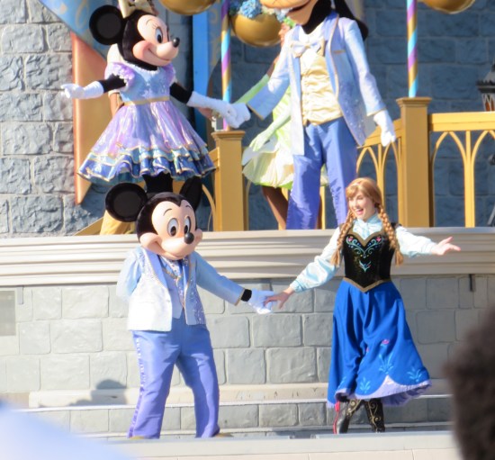 Cast members dressed as Mickey Mouse in a suit and Anna from "Frozen" hold hands on a stage. On a platform above stand Minnie and Goofy, dressed in the same royal shades of light blue and purple.