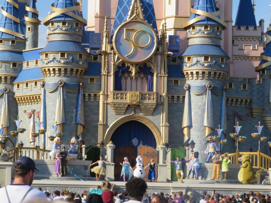 Cinderella Castle facade with a giant 50 insignia hung above a long stage with at least 14 Disney performers dancing or bouncing.