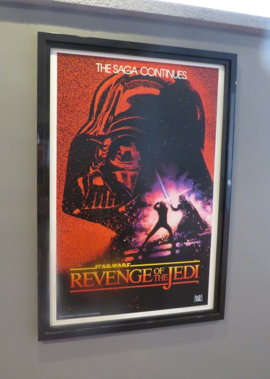 1982 "Revenge of the Jedi" poster, Vader's black head on a red background. Superimposed is a smaller image of him lightsaber-dueling Luke Skywalker. "The Saga Continues" it says on top.