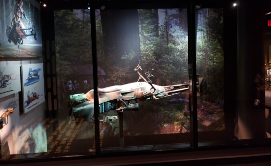 Actual speeder bike exhibited behind glass with an Endor-like forest backdrop and little Imperial blast-shield bunker in one corner.