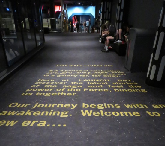 All-black gallery entrance bears a paragraph on the floor in yellow and angled like a Star Wars intro crawl: Star Wars Launch Bay. It is a period of celebration. The Star Wars saga lives on with new adventures set in a galaxy far, far away. Here at Launch Bay, discover the latest stories of the saga and feel the power of the Force, binding us together. Our journey begins with an awakening. Welcome to a new era..."