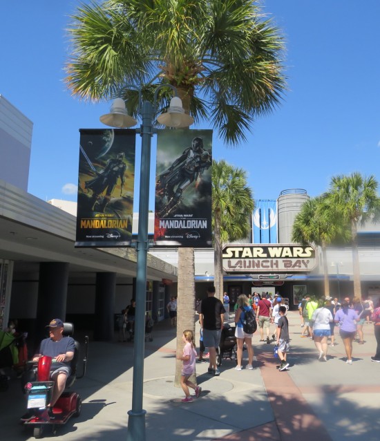 Two banners advertising "The Mandalorian" hang from a street light. In the background is the Star Wars Launch Bay building. Palm trees taller than the street lights line the sidewalk.