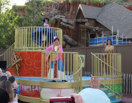 Two-tier parade float with Mirabel Madrigal twirling on top, Mulan and Pocahontas waiving to the crowd from the lower level.