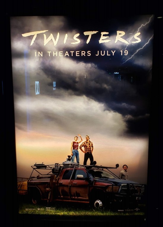 Twisters movie poster shows release date of July 19th above the two leads standing on a red ruck and looking at an imminently stormy sky. 