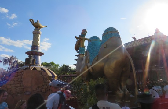 Disney's Aladdin theme-park section with genie lamp atop a tall column and a camel statue facing away from us. Both are shadowed because the sun is pointing into the camera.