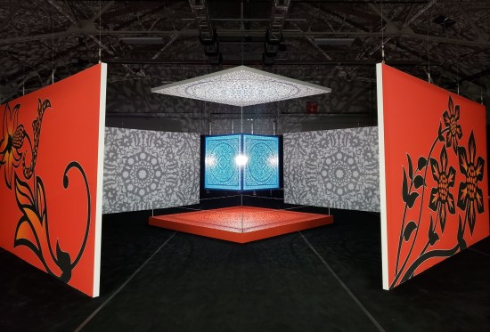Blue metal cube hanging from ceiling and projecting intricate shadows onto surrounding platforms and walls.