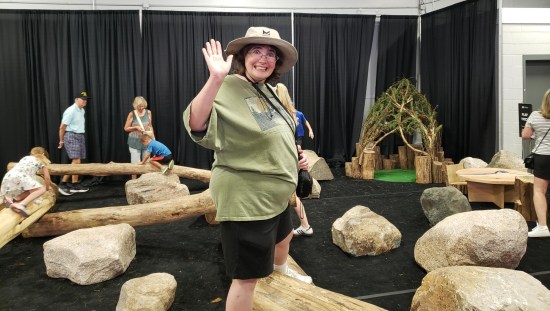 In an indoor playground made solely of logs and fake boulders, Anne stands on a log and waves enthusiastically at the camera. 