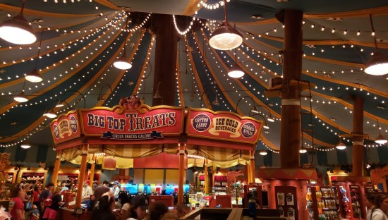Inside a brightly lit building shaped like a circus tent filled with concession stands.