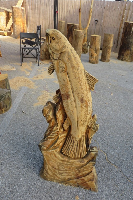 Big fish, like a trout or bass or whatever, carved from a log and standing on its tail fins.