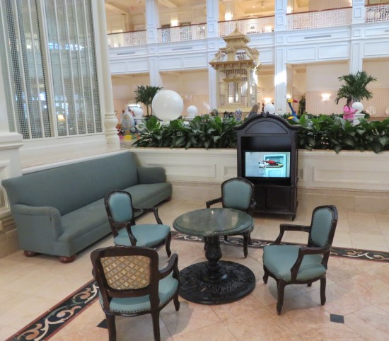 Unwalled lounge with four blue child-sized dining-room chairs, a blue child-sized couch, and a child-sized cabinet with a child-sized TV showing a Goofy cartoon.