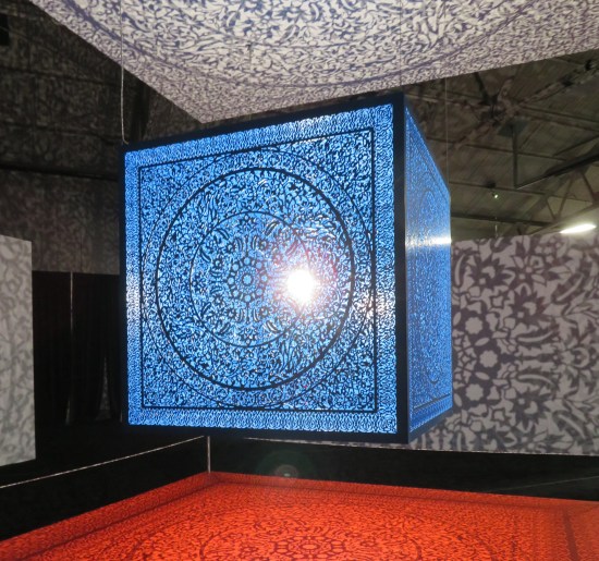 Blue cube suspended in midair with a light in the middle of it. Each metallic cube side is intricately patterned to project shadows on the surfaces surrounding it.