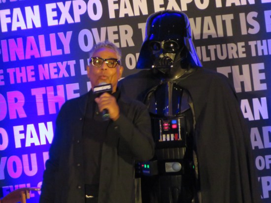 Giancarlo Esposito talking onstage with a Darth Vader cosplayer standing to one side behind him.