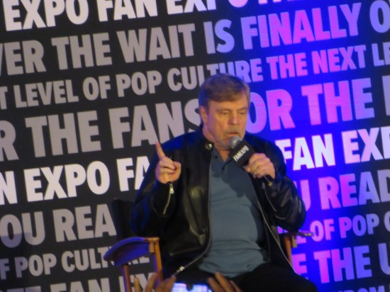 Mark Hamill seated onstage, talking and holding up his right index finger.