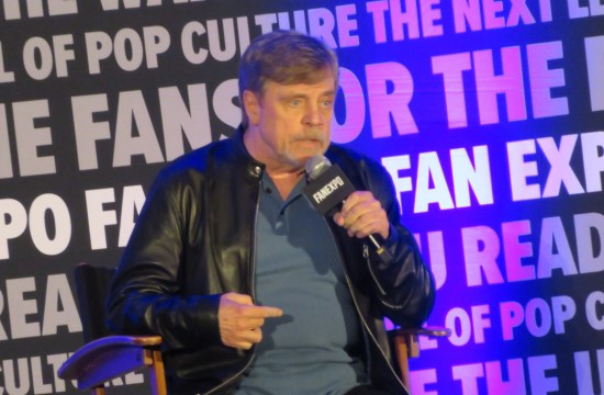 Mark Hamill sitting onstage, talking into a mic and pointing at his stomach.