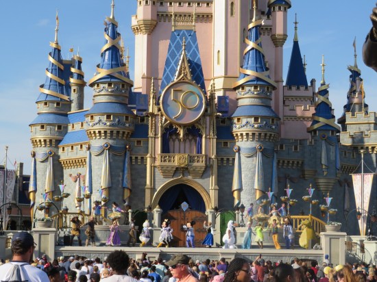 Cinderella Castle shot closely enough to cut off its spires but far enough away that the dozen or so cast members dancing onstage are tiny. A giant Disney World 50th-anniversary seal is still mounted on the castle.