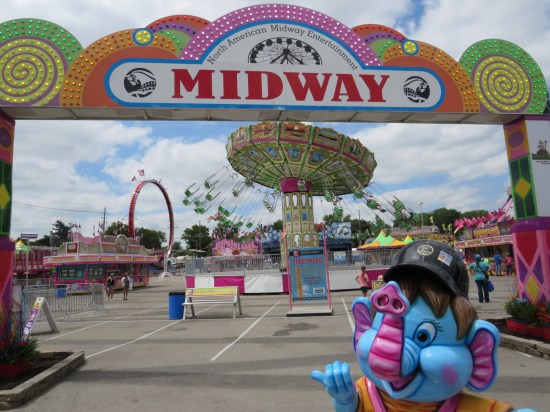 State fair carnival rides with a big Midway sign and a cutesy blue elephant mascot statue welcoming guests.